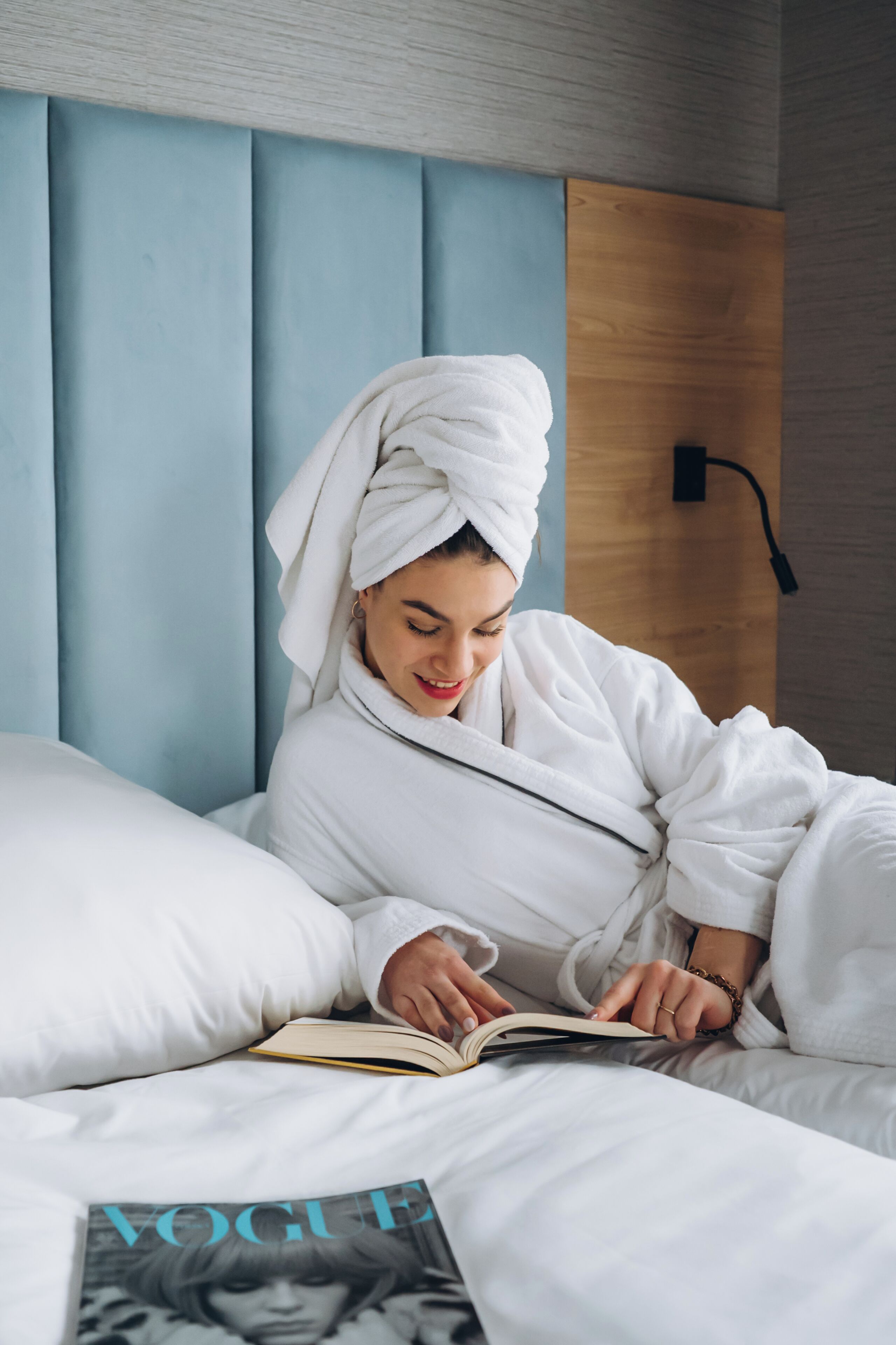 A person in a white bathrobe reading a book on a bed with a blue headboard and a wooden panel.