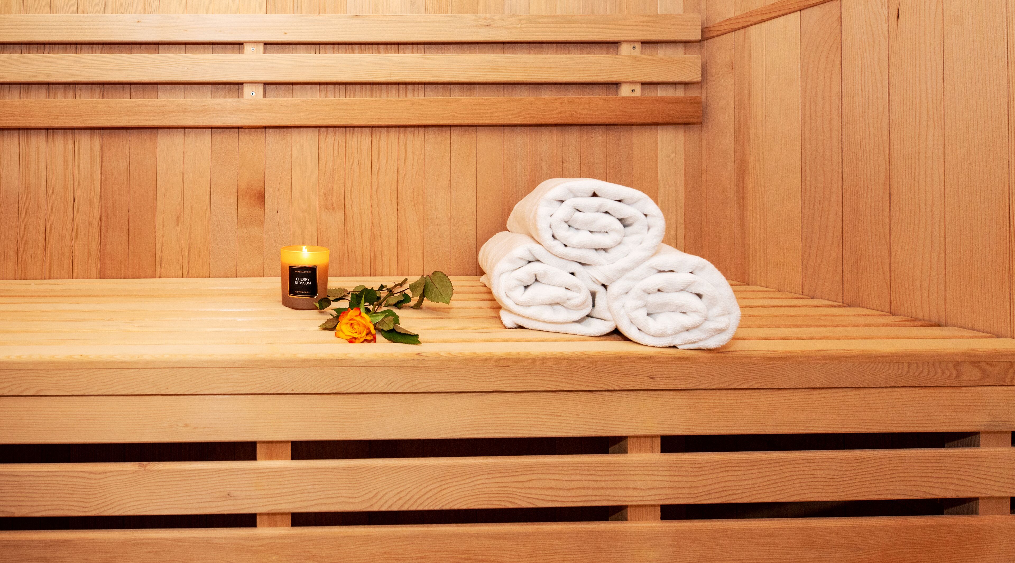 A wooden sauna bench with rolled towels and a lit candle.