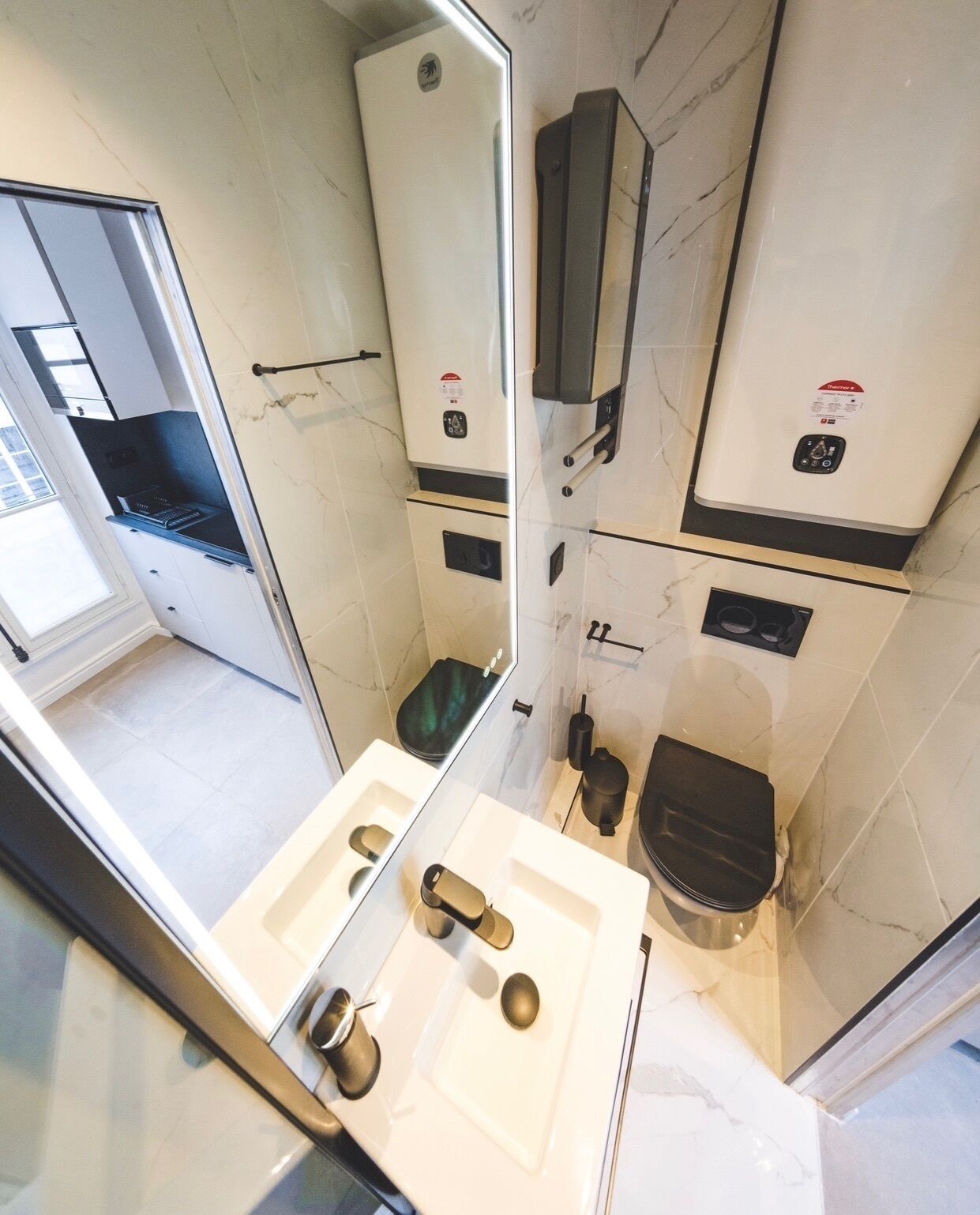 Een kleine badkamer met een wastafel, toilet en spiegel. Een handdoekhouder zit aan de muur bevestigd, en een klein raam zorgt voor natuurlijk licht.