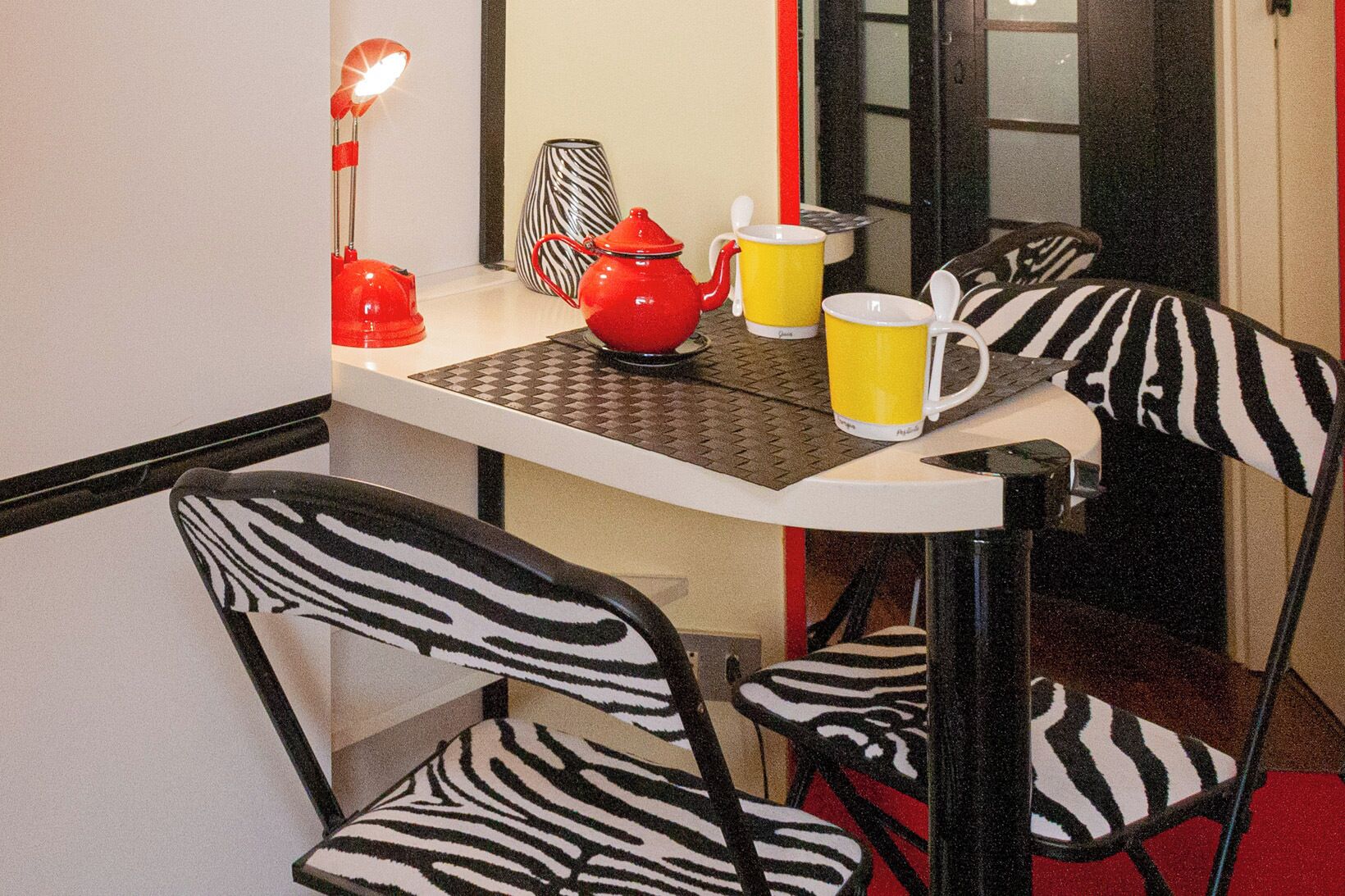 A dining area with zebra-striped chairs, a table with a zebra-patterned placemat, a red teapot, yellow mugs, and a red lamp.