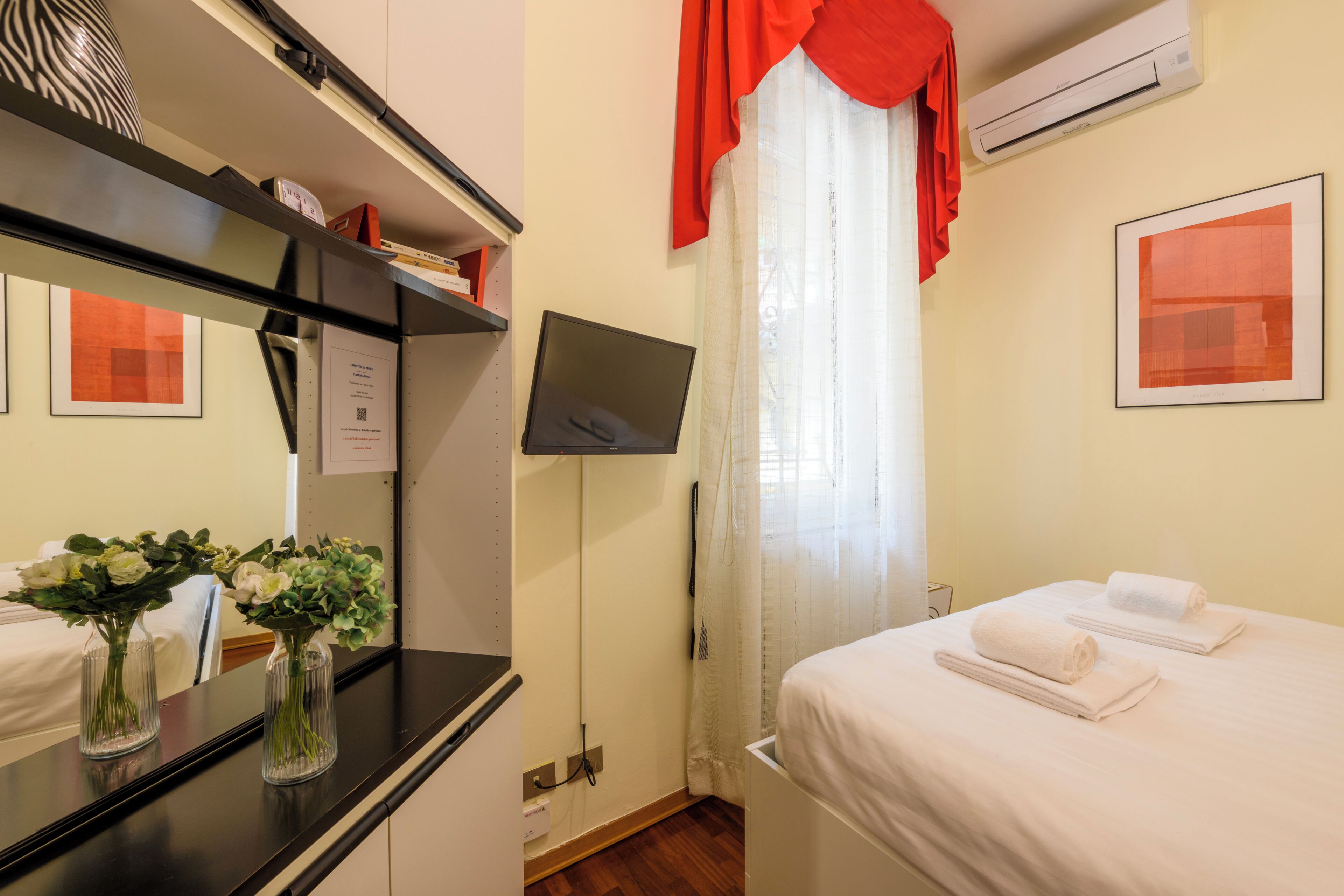 Una habitación de hotel con cama, televisión, espejo y escritorio con un florero.