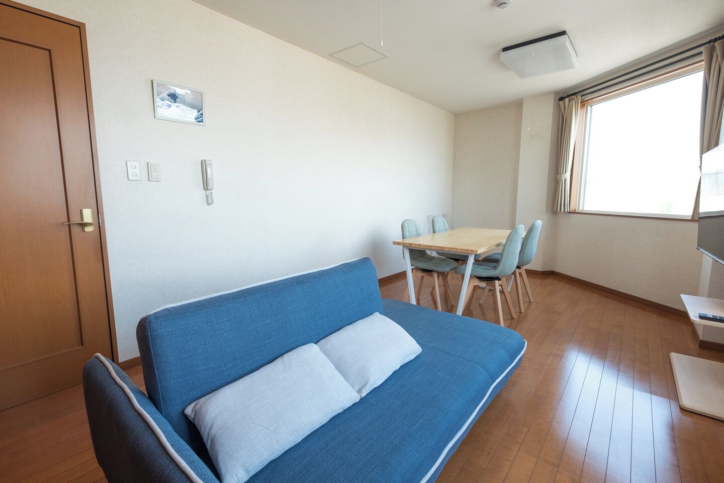 Ein Zimmer mit einem blauen Sofa, einem Holzesstisch mit Stühlen, einem kleinen Fernseher und einem Fenster mit Vorhängen.