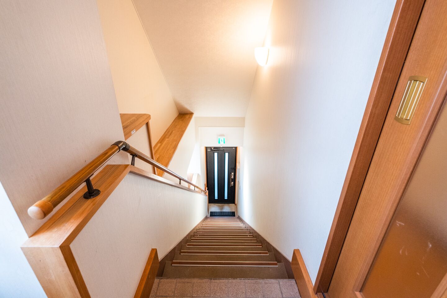A modern staircase with wooden handrails and a carpeted floor leading to a door with a green exit sign.