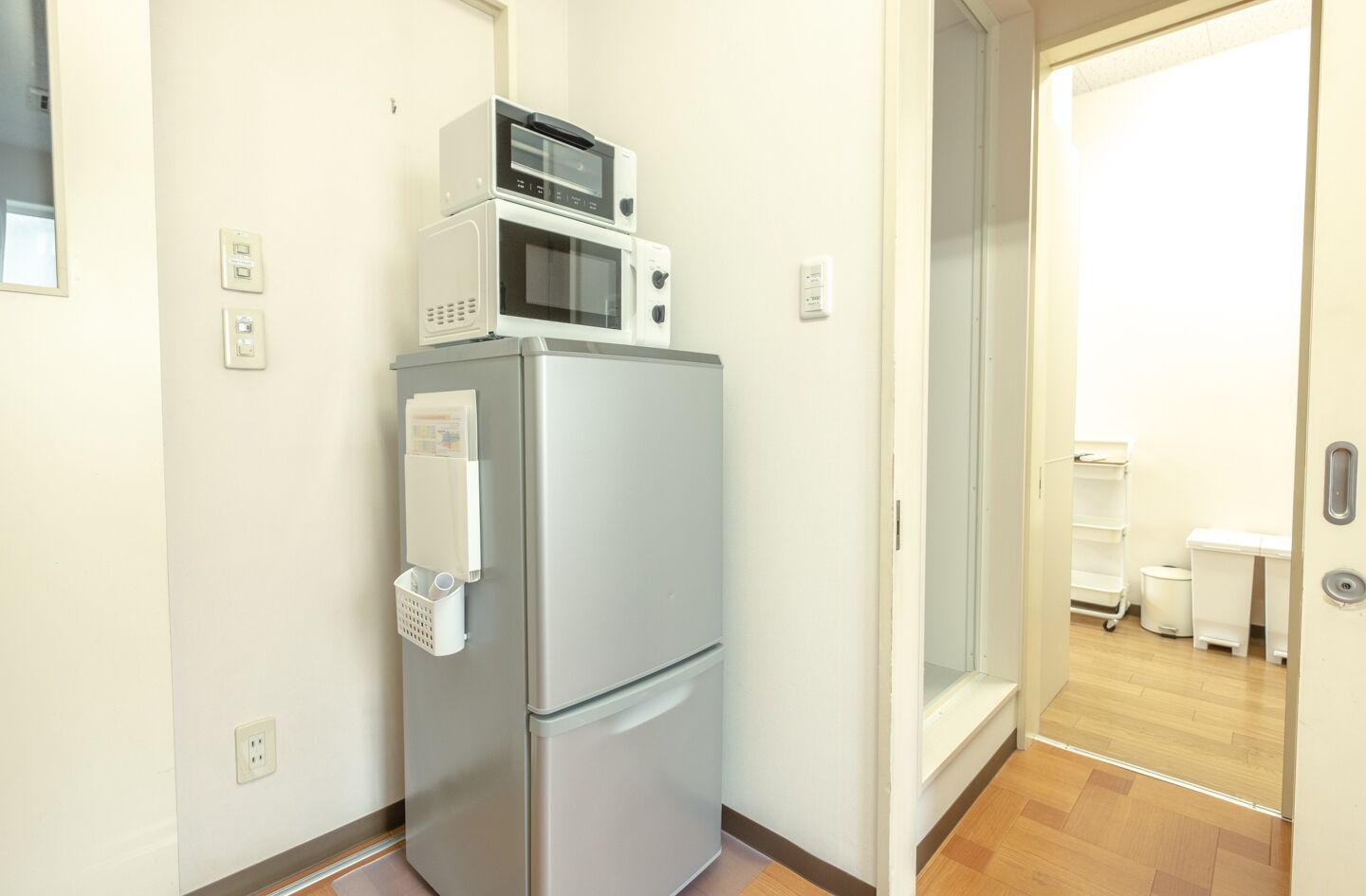 A compact kitchen with a refrigerator, microwave, and a small freezer unit. There is a white shelf unit with a basket and a paper holder. The room has a wooden floor and a door leading to another area.