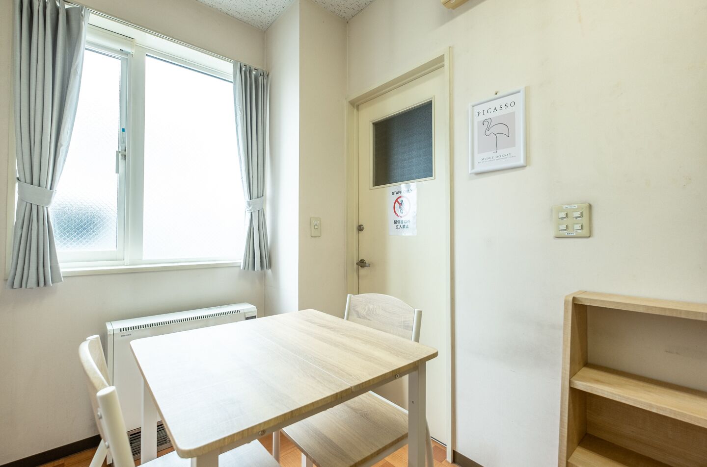 A room with a wooden table, white chairs, a radiator, a window with curtains, a door with a sign, and a wall-mounted light switch.