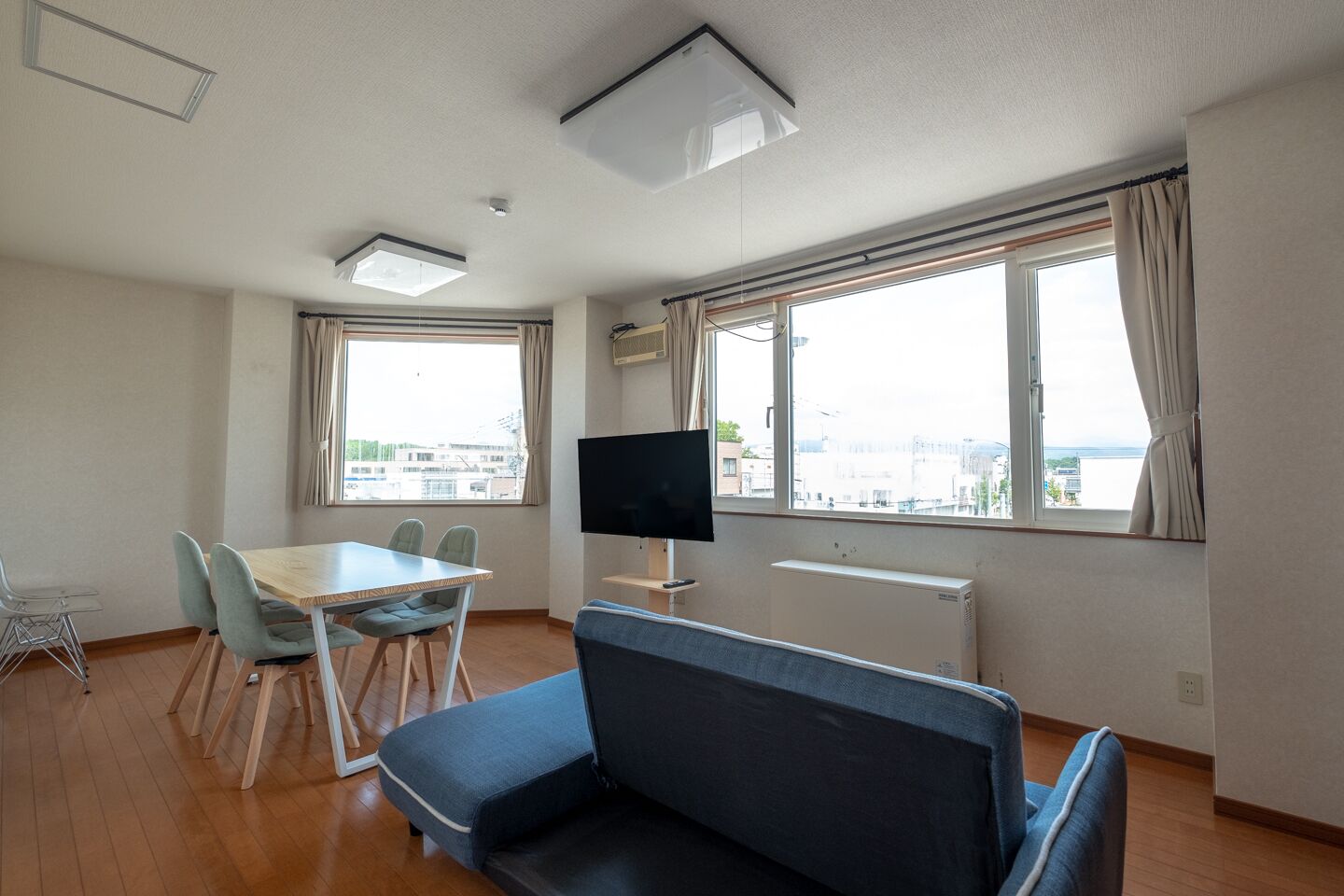 A modern room with a dining table, chairs, a sofa, a TV, and large windows offering a city view.