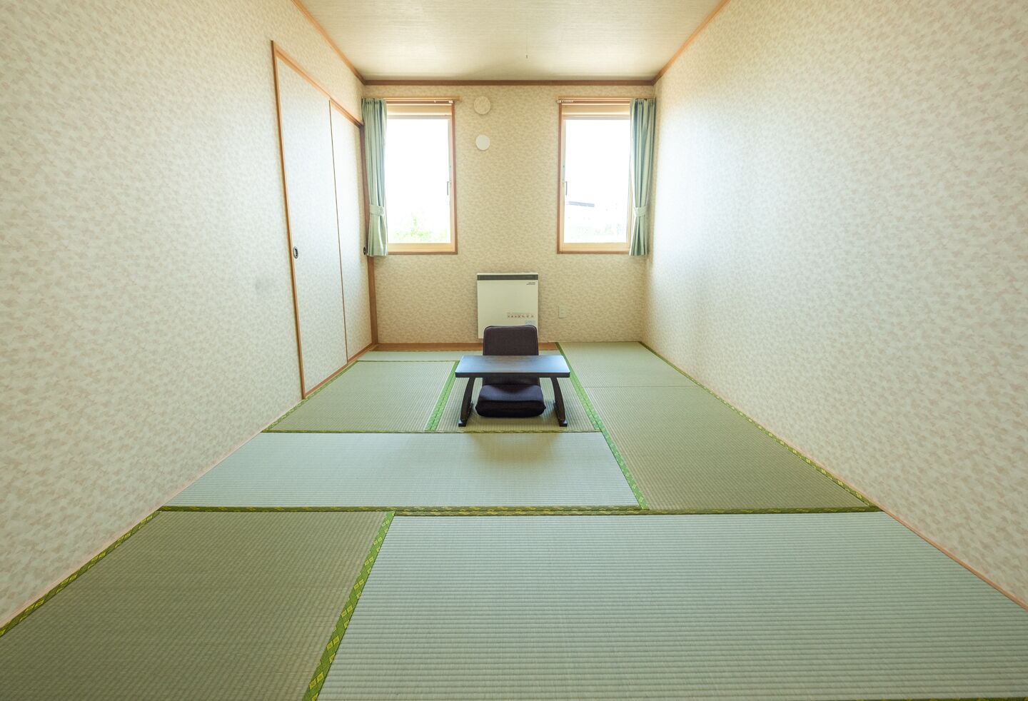 A room with tatami mat flooring, a desk with a chair, two windows with curtains, and a cabinet.