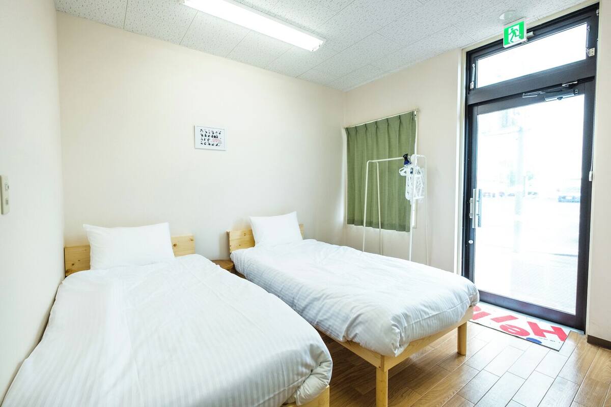 A hotel room with two single beds, a window with a curtain, a green exit sign, and a door with a handle.