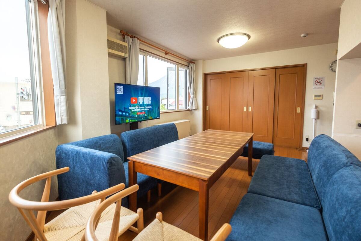 A room with a wooden table and blue upholstered chairs, a television on the wall, and a large window with curtains.