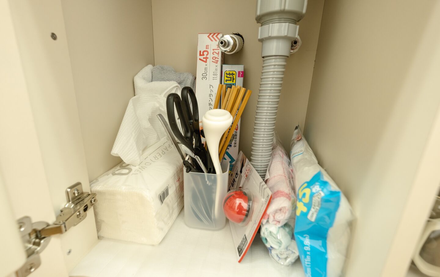 Un petit espace de rangement avec une étagère blanche contenant un sac en plastique blanc, un récipient en métal avec des ciseaux à manche noir, un récipient en plastique blanc avec des outils à manche jaune, un récipient en plastique blanc contenant un objet rouge et un sac en plastique bleu.