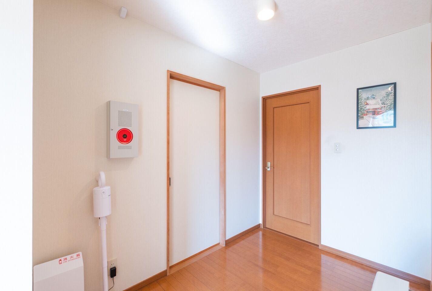 A hallway with wooden flooring, a white wall-mounted fire alarm, a door, and a framed picture.