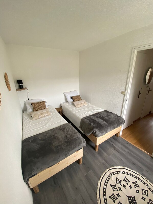 A room with two single beds, a small bedside table with a lamp, a wall-mounted shelf, and a wooden floor. - Chaleureuse Maison à la Plage (Courseulles-sur-Mer)
