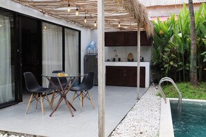 Un área de comedor al aire libre con una mesa y sillas de vidrio, una cocina con armarios de madera y una piscina con una escalera de metal.