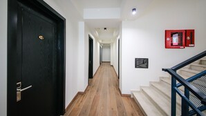 A hallway with a black door, wooden flooring, a staircase with blue railings, and a red fire alarm on the wall.