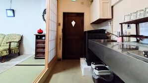 Cocina pequeña con encimera negra, un lavamanos y un armario arriba. Se ve una puerta con un adorno blanco en forma de diamante, un estante con un jarrón rojo, y una silla al fondo.