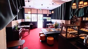 Un interior de restaurante con alfombra roja, mesas de madera, taburetes naranjas, lámparas colgantes y una vitrina con pasteles.