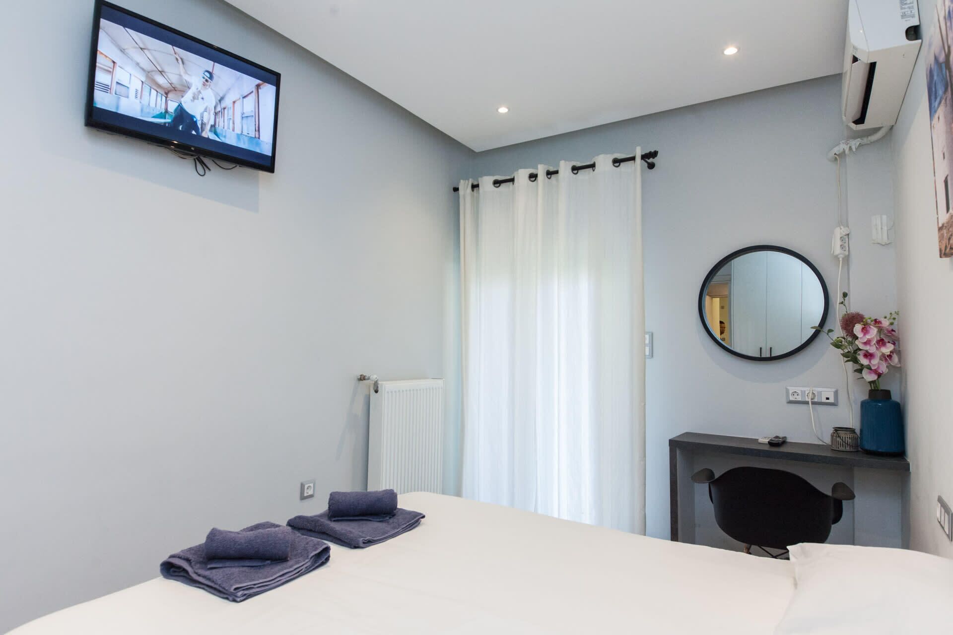 A hotel room with a bed, a desk, a chair, a mirror, a TV, and a radiator.