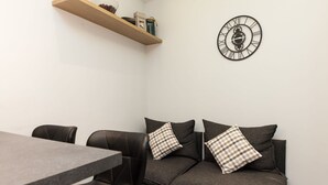 Een moderne woonkamer met een grijze bank, twee geruite kussens, een houten plank met decoratieve objecten en een wandklok.