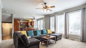 Ein Wohnzimmer mit einem Ecksofa, einem Couchtisch und einem Deckenventilator. Im Hintergrund ist eine Küche mit Holzschränken und einer Mikrowelle zu sehen.