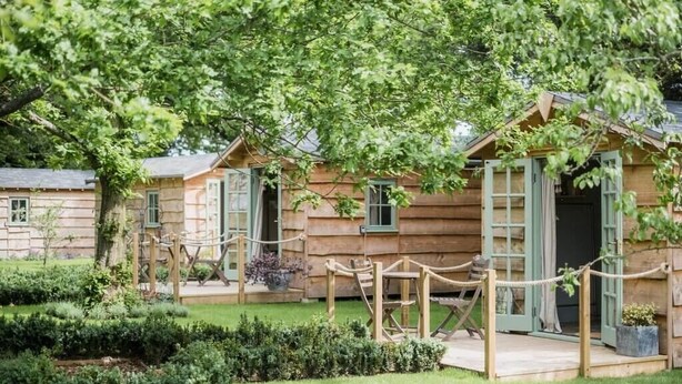 Una hilera de cabañas de estilo rústico con contraventanas verdes, rodeadas de jardines cuidados y una barrera de cuerda.