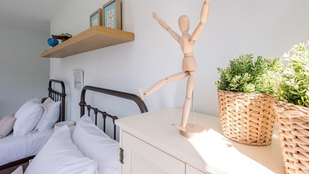 A bedroom with a bed, a wooden mannequin, a bedside table, a wicker basket, and a wall-mounted shelf with pictures. - Springfield, Westleton (Saxmundham)