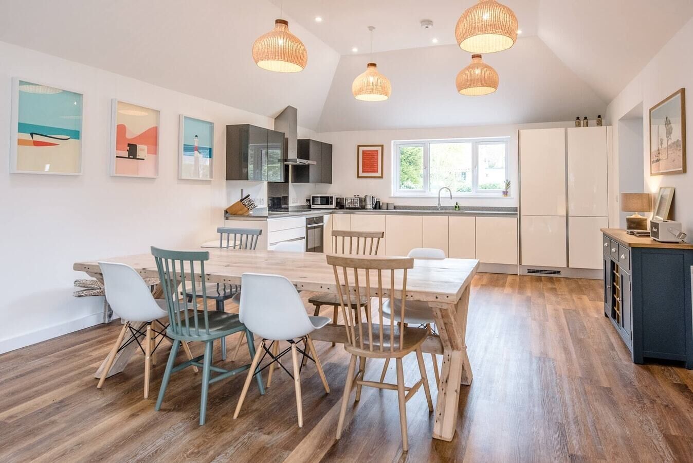 Une cuisine moderne avec des meubles blancs, une table à manger en bois et des chaises. Des luminaires suspendus sont placés au-dessus de la table.