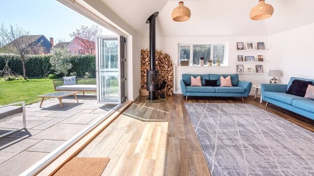 A modern living room with a large glass door leading to a patio, a blue sofa, a fireplace, and a view of the garden. - Springfield, Westleton (Saxmundham)
