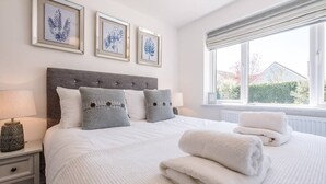 A neatly made bed with white linens and two decorative pillows, a bedside table with a lamp, and a window with a view of the outside. - Springfield, Westleton (Saxmundham)