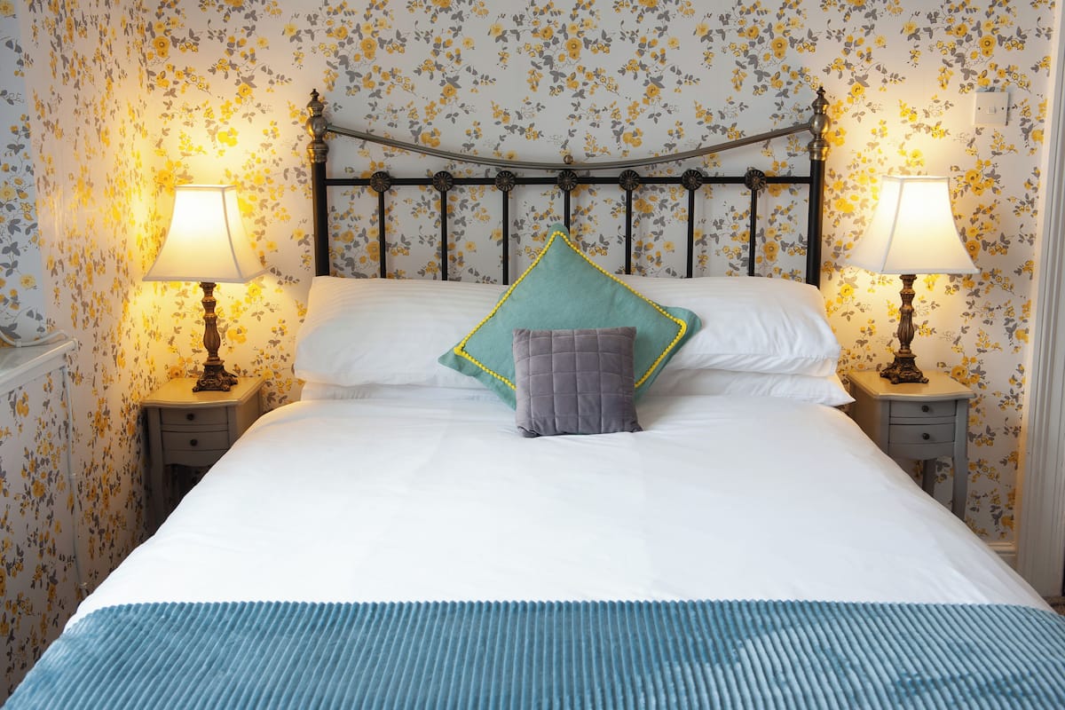 A neatly made bed with a blue bedspread, two nightstands with lamps, and floral wallpaper.