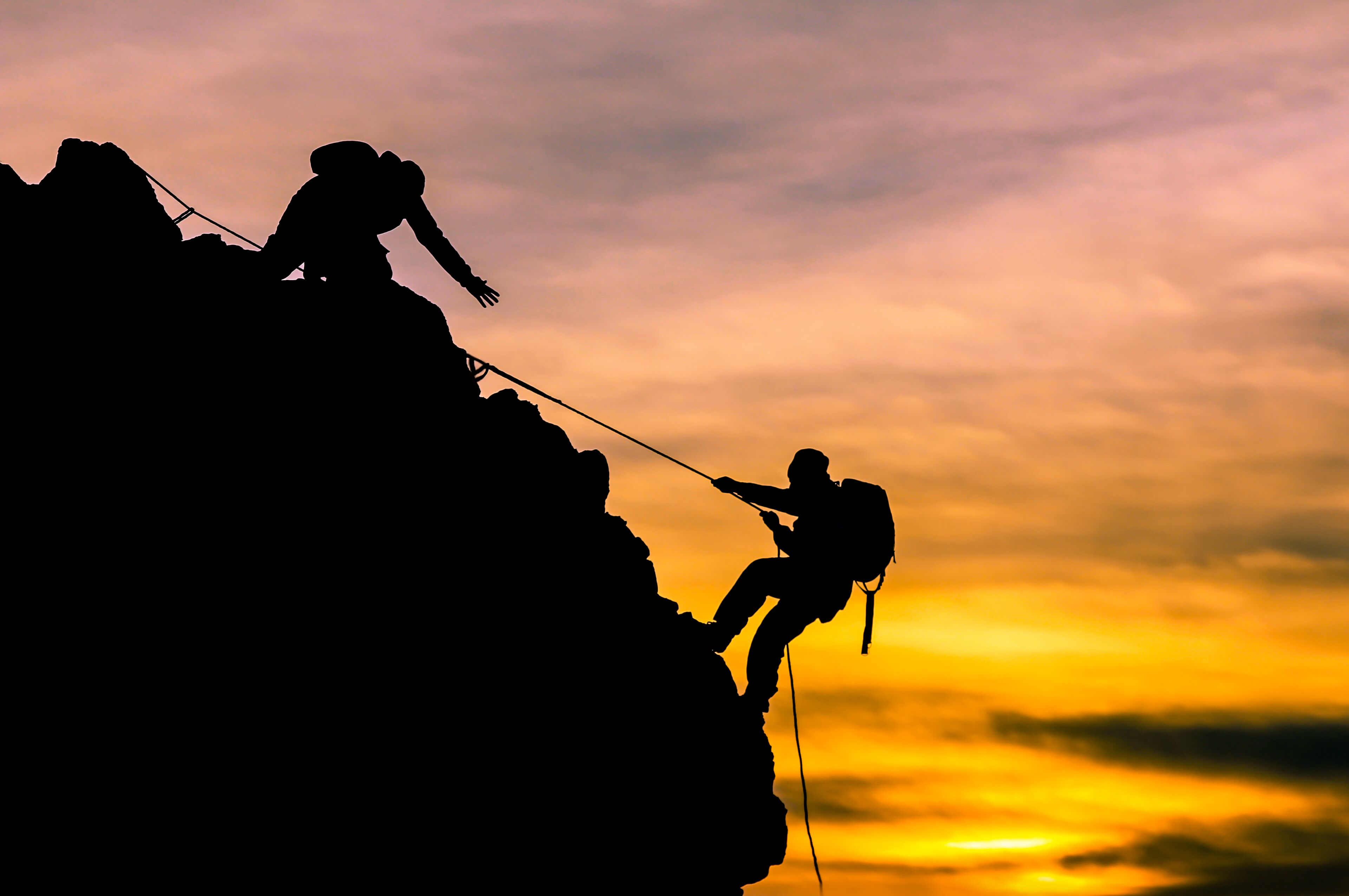 Dos figuras recortadas escalando una montaña con un atardecer de fondo.