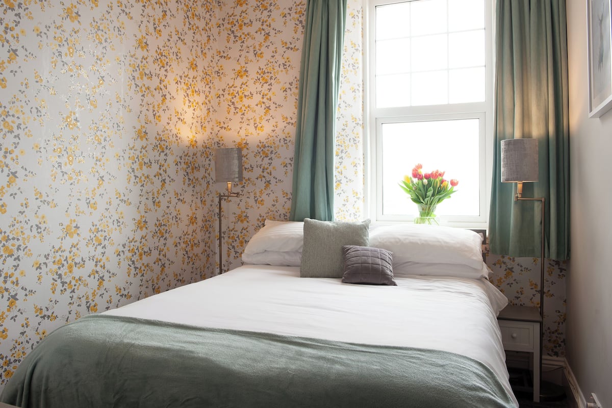 A neatly made bed with a green bedspread, floral wallpaper, and a window with green curtains.