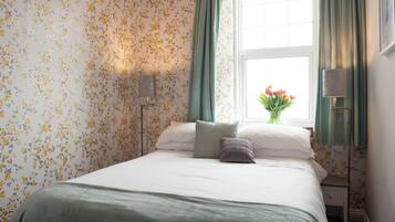 Un lit bien fait, recouvert d’une couvre-lit verte, avec un papier peint à motifs floraux et une fenêtre ornée de rideaux verts.