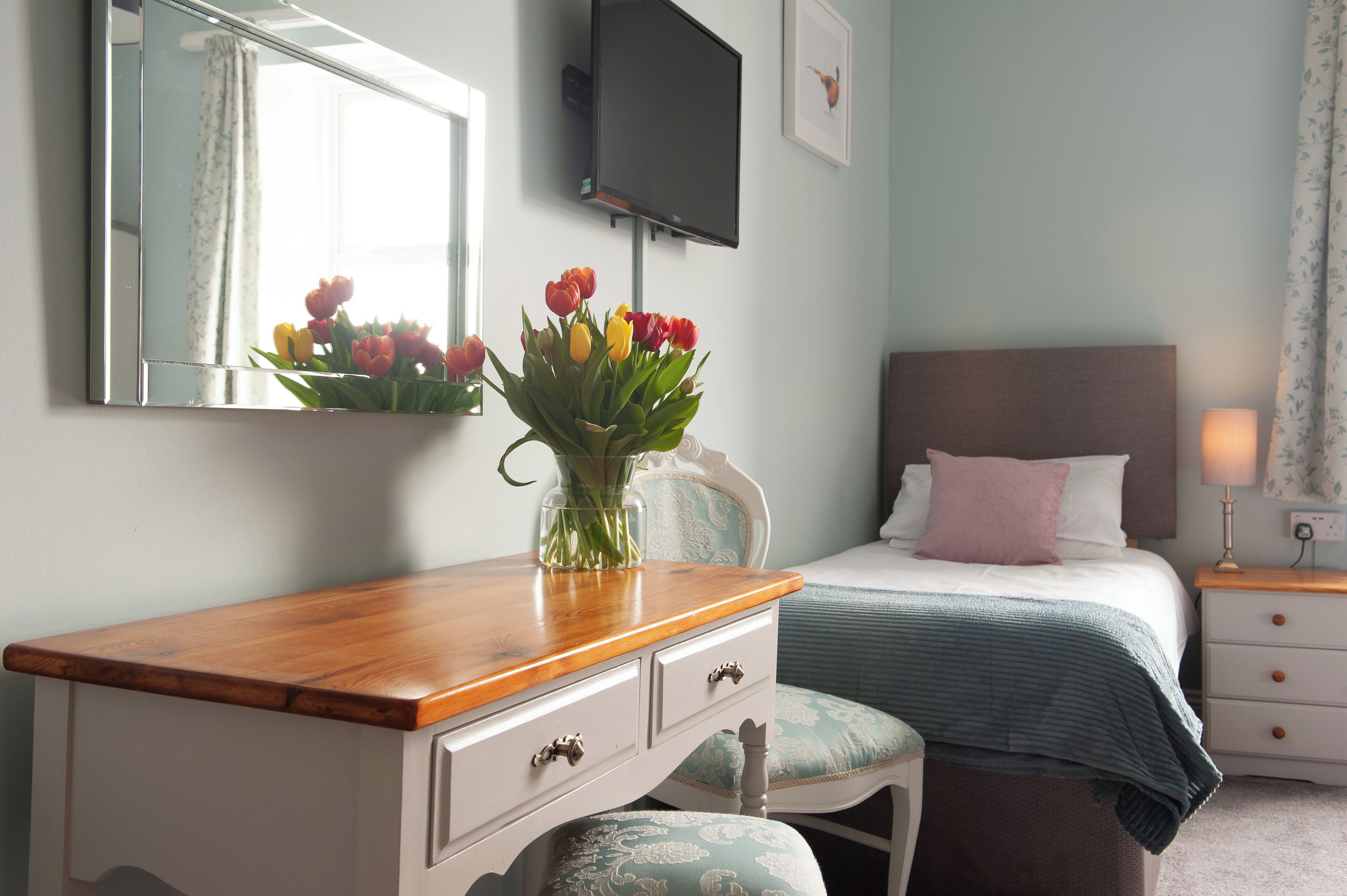 A bedroom with a wooden dresser, a bed with a blue and white bedspread, a nightstand with a lamp, and a wall-mounted TV.