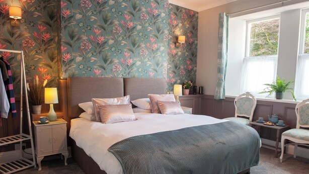 A bedroom with a bed, nightstands, a dresser, a chair, a window with curtains, and a floral wallpaper.