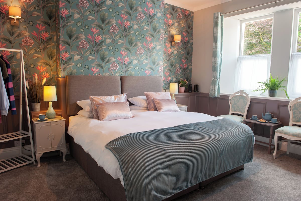 A bedroom with a bed, nightstands, a dresser, a chair, a window with curtains, and a floral wallpaper.