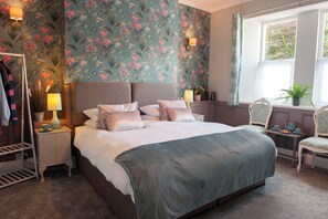A bedroom with a bed, nightstands, a dresser, a chair, a window with curtains, and a floral wallpaper. - Duchy House B&B (Yelverton)