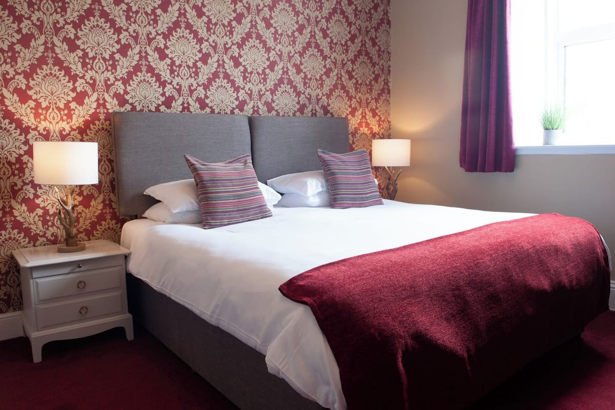 A neatly made bed with white linens and two pillows, a red throw blanket, and two bedside lamps on a nightstand.