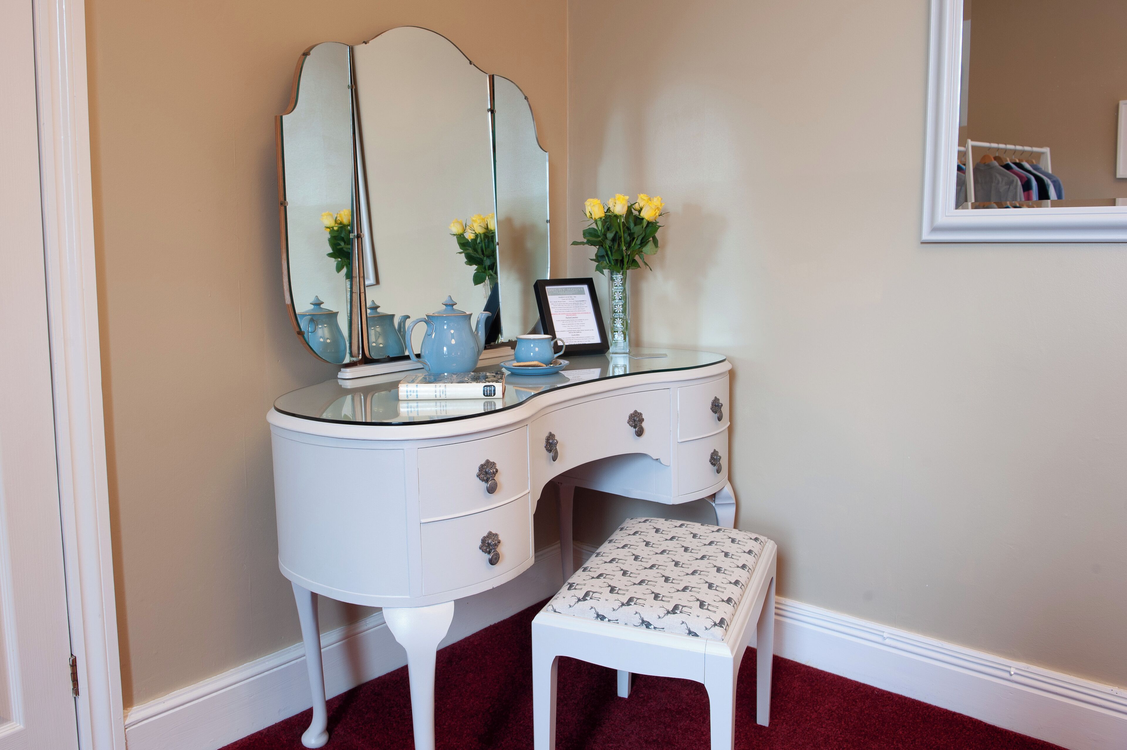 A white vanity with a large mirror, a blue teapot, a cup, and a vase with yellow flowers.