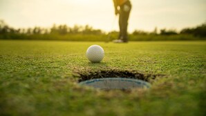 A golf ball on a green with a hole in the foreground and a golfer in the background.