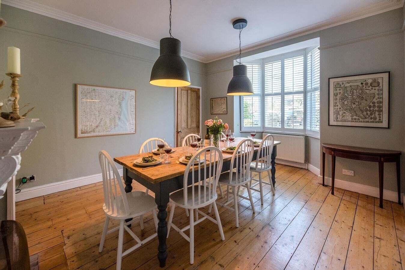 Une salle à manger avec une table en bois dressée pour six personnes, des chaises blanches, deux lampes suspendues et des œuvres d’art accrochées aux murs.