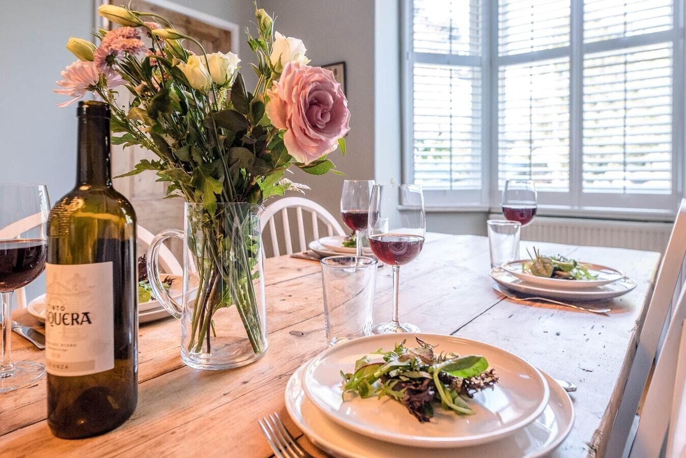 Una mesa de comedor con una botella de vino, un jarrón de flores y platos con comida y copas de vino.