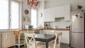 A modern kitchen with white cabinets, a stainless steel refrigerator, a dining table with chairs, a microwave, and a decorative chandelier. - BnButler - Riccardo Galli 9 (Milan)