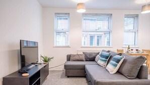 A modern living room with a grey sofa, a flat-screen TV, and a small side table. There are three windows providing natural light.