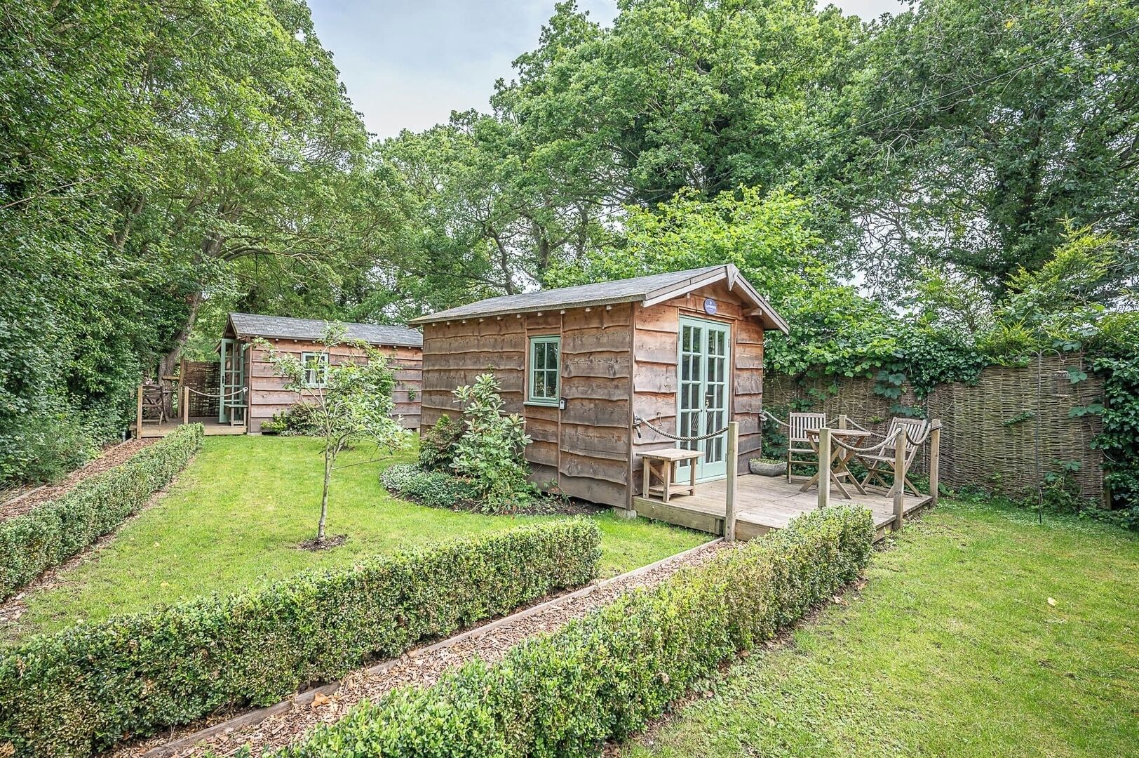Una cabaña rústica de troncos con un porche y mobiliario exterior, rodeada de un jardín cuidado y árboles.
