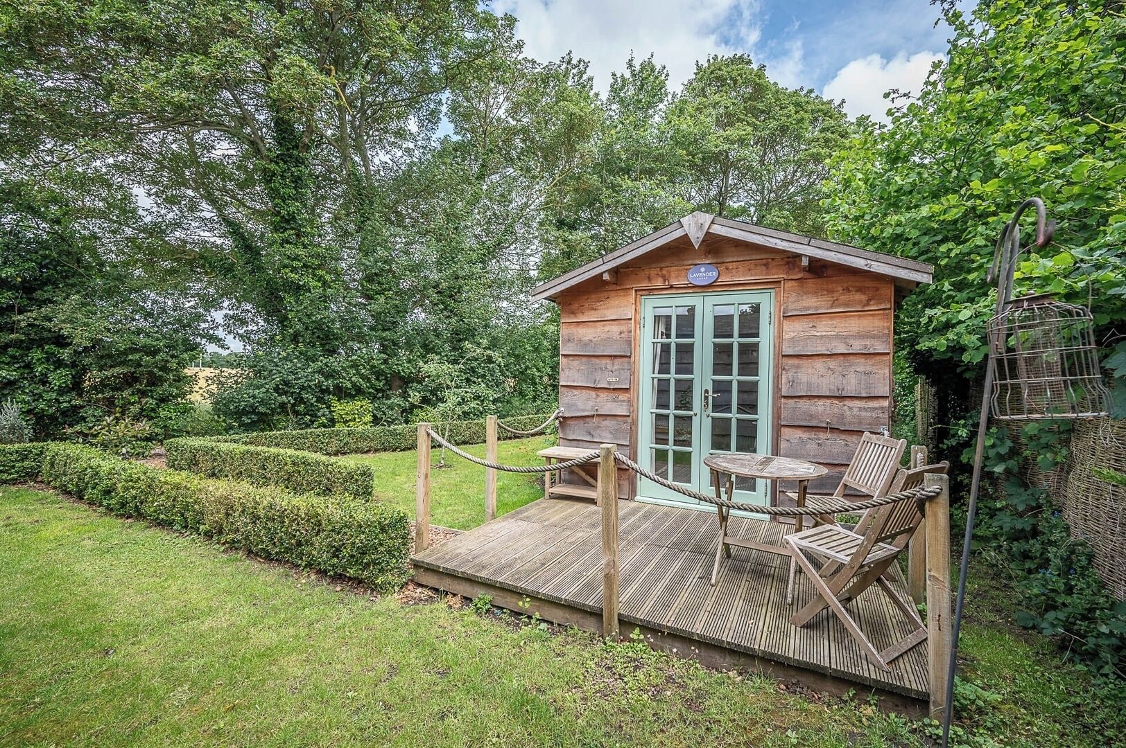 A wooden cabin with a green door, a small porch with a table and chairs, surrounded by a well-maintained garden and trees.