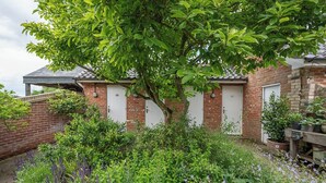 A brick building with two white doors, surrounded by lush greenery and flowering plants. - Feather Lodge, Toad Hall, Wangford (Wangford)