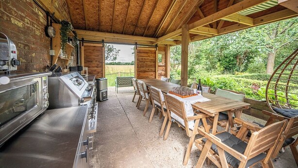 A rustic outdoor kitchen with a large stove, wooden table, and chairs, surrounded by a wooden pergola and lush greenery. - Feather Lodge, Toad Hall, Wangford (Wangford)
