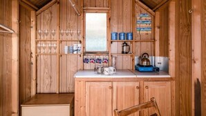 A wooden cabin kitchen with a wooden counter, shelves with glassware, and a kettle. - Driftwood Beach Hut, Southwold (Suffolk)