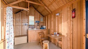 A wooden cabin with a kitchen area, a dining table, and a small stool. - Driftwood Beach Hut, Southwold (Suffolk)