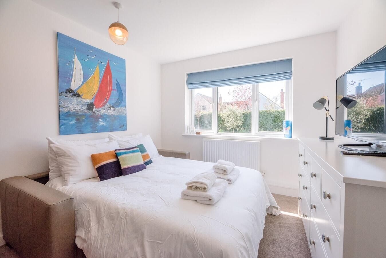 A bedroom with a bed, a painting of sailboats, a window with a view, and a desk with a lamp.